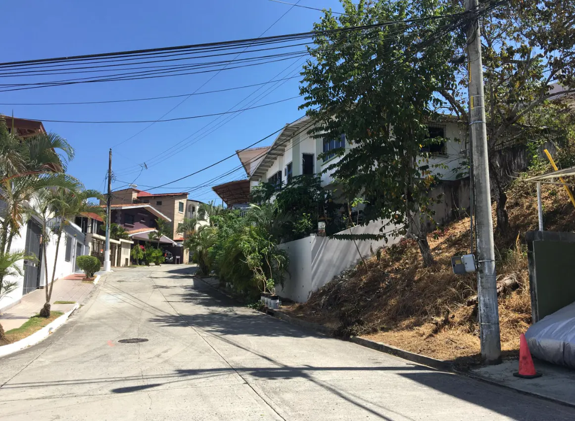 A street view with houses on a hill under a clear blue sky. Power lines are visible above the street.