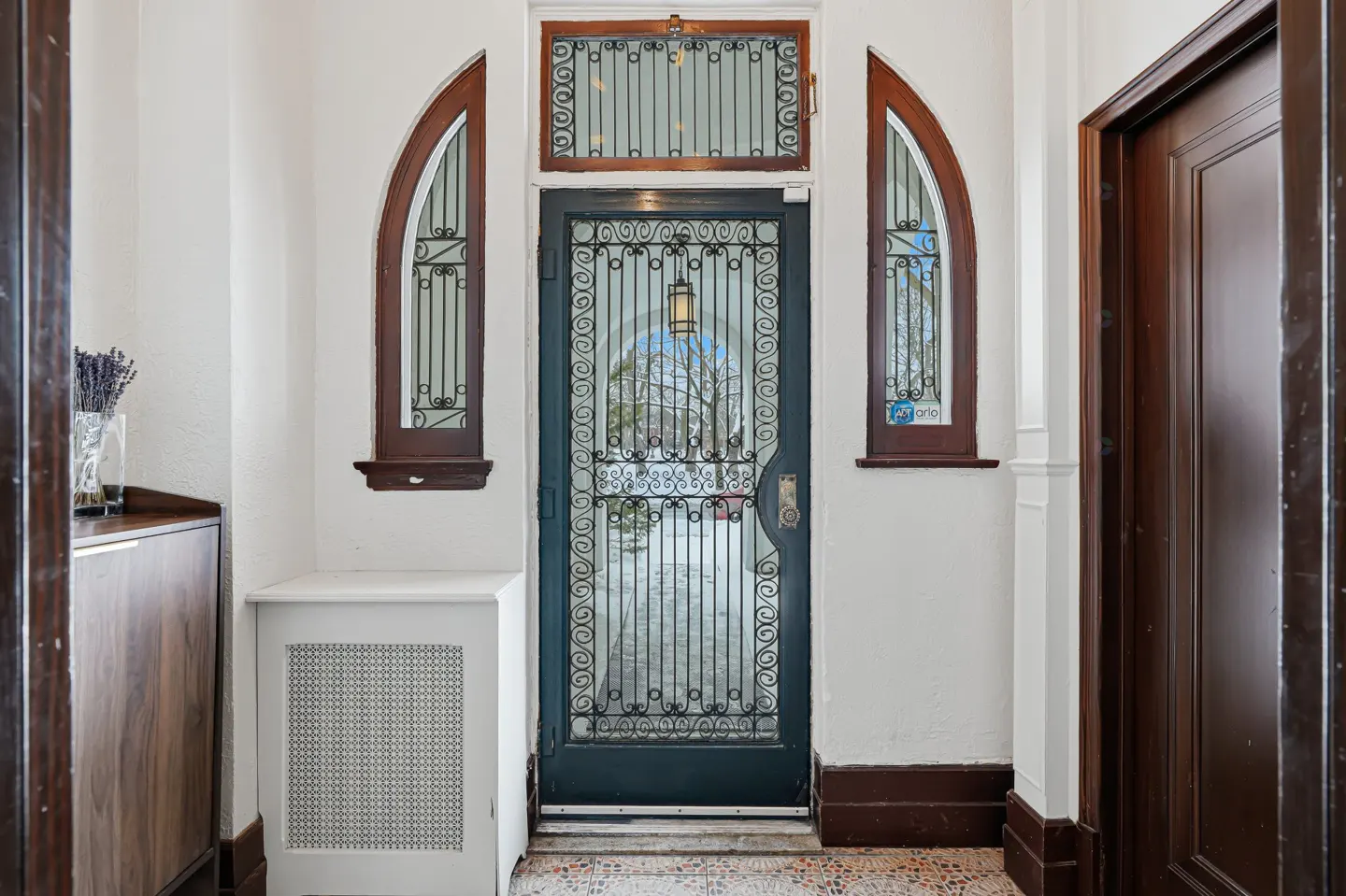 Foyer with a dark blue door with decorative ironwork, flanked by arched windows. A wooden cabinet and radiator cover are on the left.