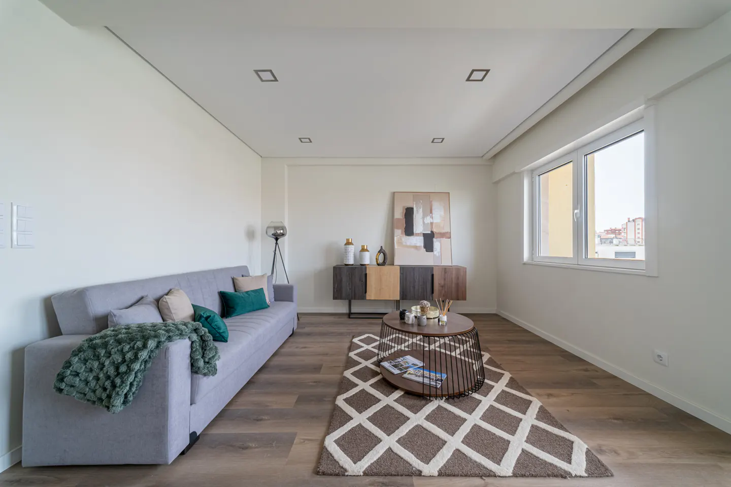 Living room with a gray sofa, green throw blanket, and pillows. A coffee table sits on a patterned rug, with a sideboard and artwork in the background.