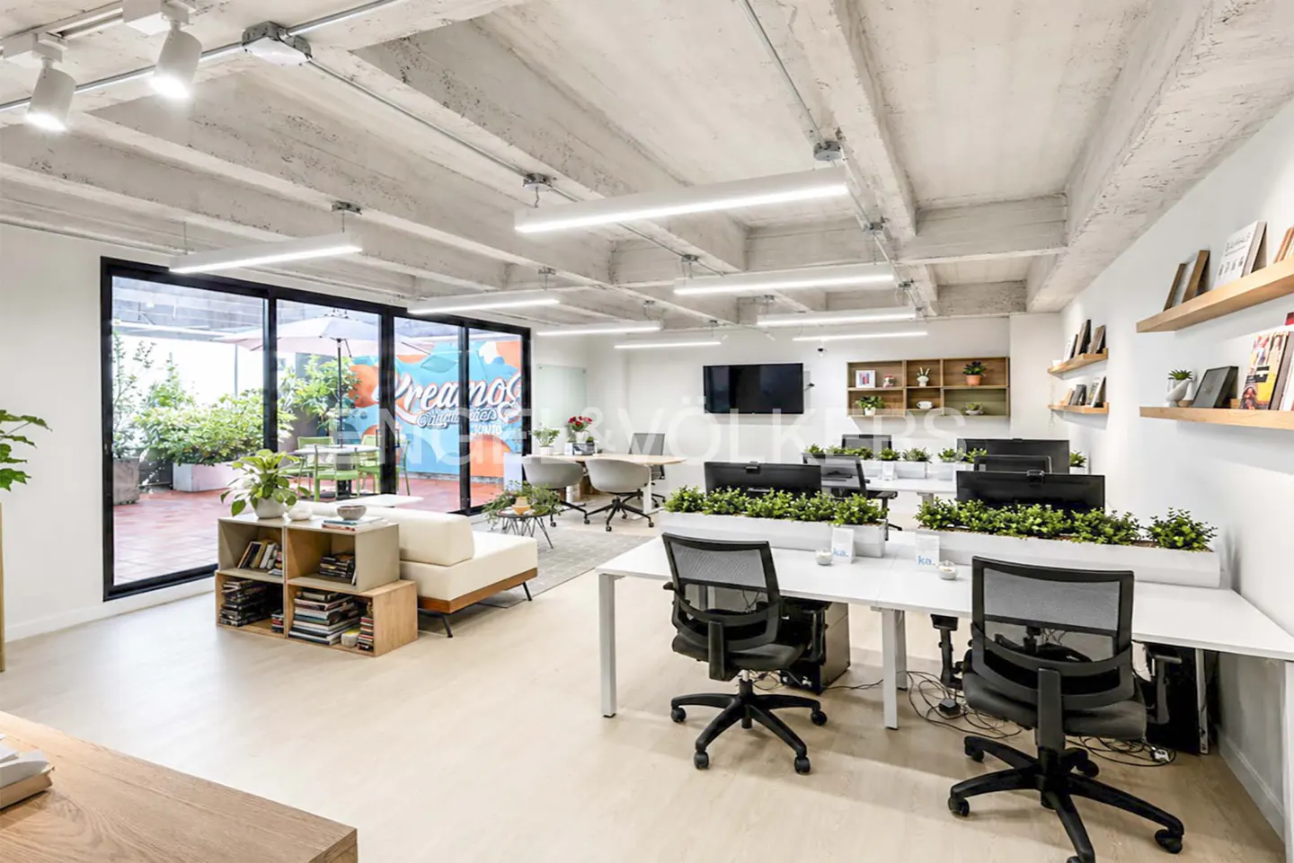 Bright, modern office space with white desks, black chairs, and greenery. A lounge area with a sofa and outdoor patio is visible.