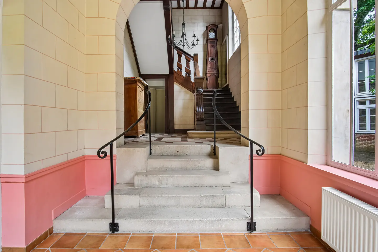 Stone steps with black iron railings lead to a grand staircase, grandfather clock, and chandelier in a tiled foyer.