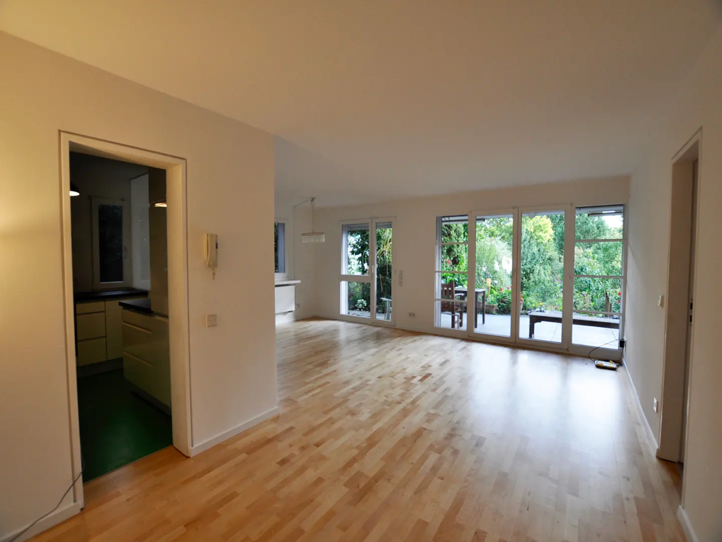 Bright, open-plan living space with light wood floors and white walls. Sliding glass doors lead to a patio with greenery. Kitchen visible through doorway.