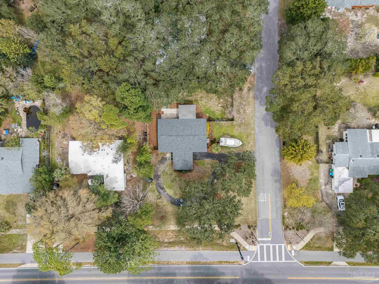 Aerial view of a gray-roofed house surrounded by trees, with a boat parked in the yard. A road runs along the bottom of the frame.