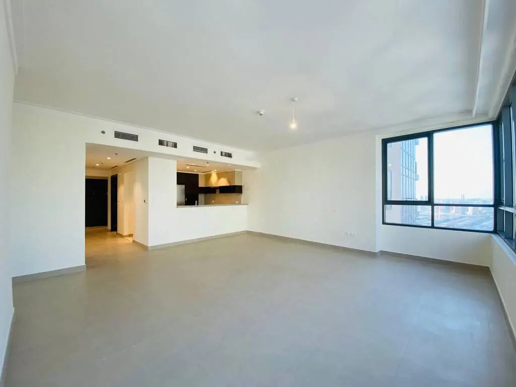 Bright, empty apartment with white walls, gray tile floor, and large window with city view. Kitchen area visible in the background.