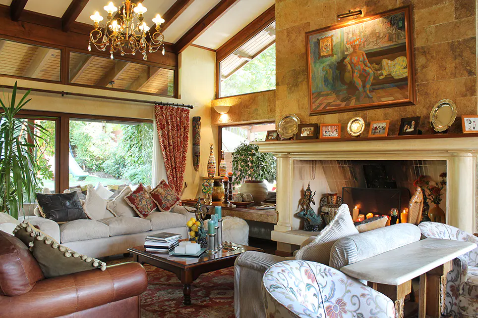 Living room with high ceilings, exposed beams, chandelier, stone fireplace, and large windows overlooking a green garden.