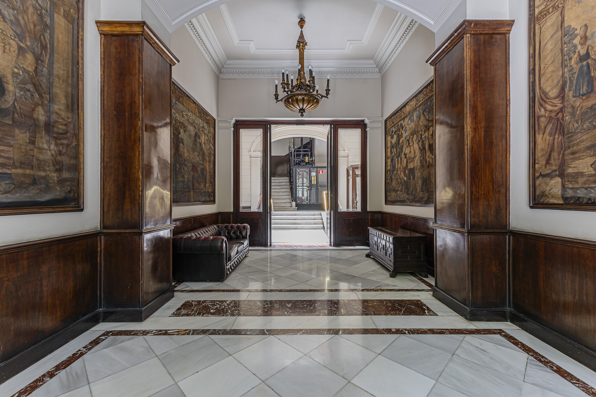 Elegant hallway with marble floors, dark wood paneling, ornate tapestries, a chandelier, and a leather sofa.