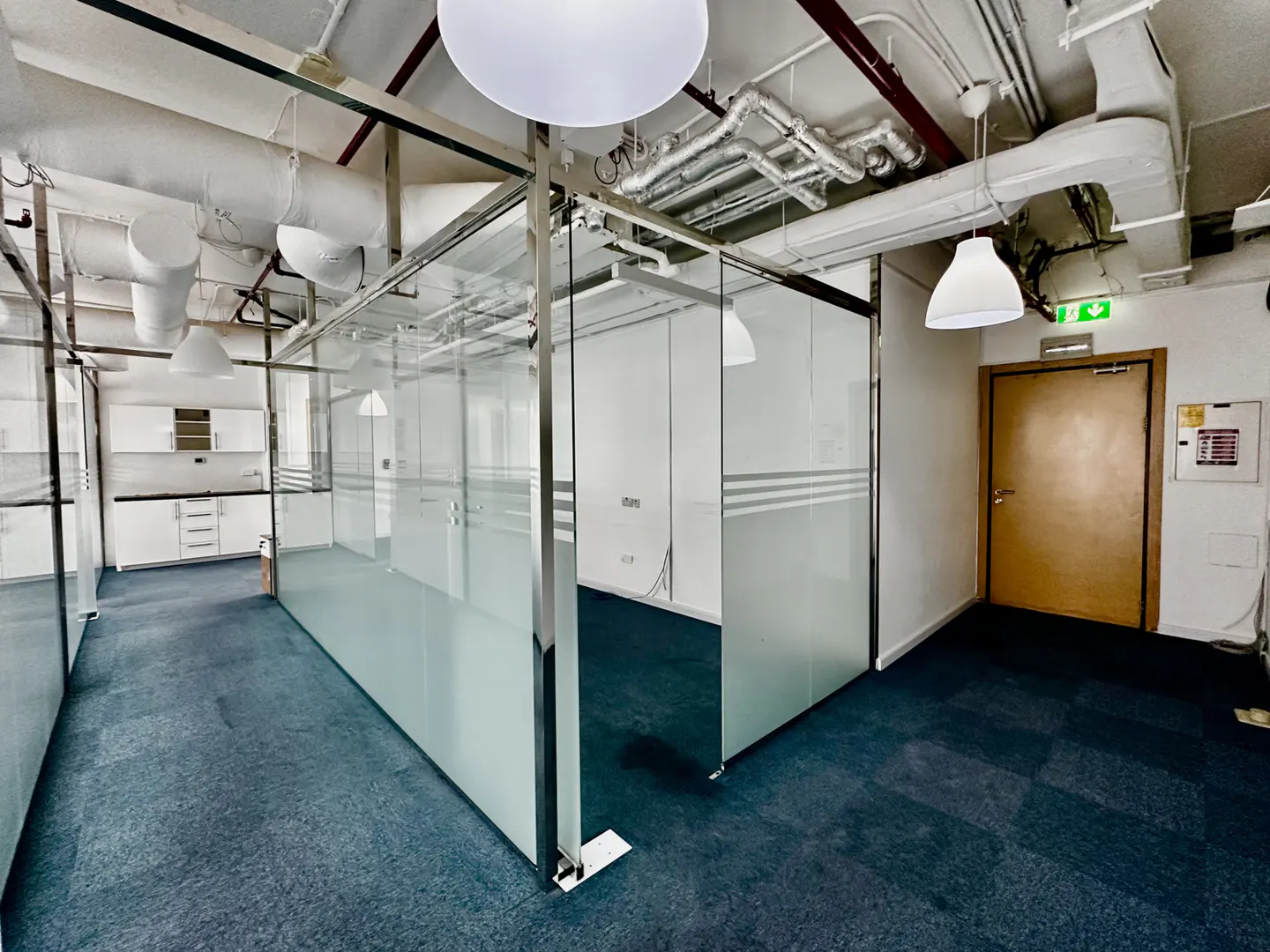 Interior view of an office space with glass-walled rooms, blue carpet, and white kitchen area in the background. Overhead lighting and exposed ductwork are visible.