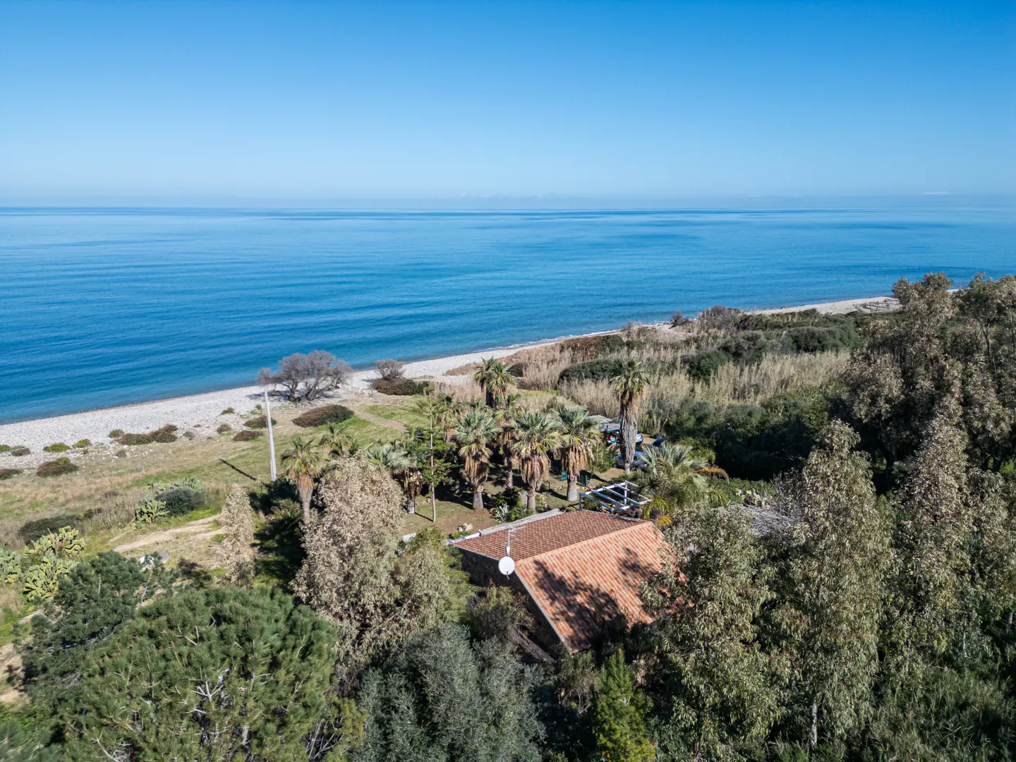 Aerial view of a house with a red tile roof surrounded by trees, near a beach and the blue ocean.