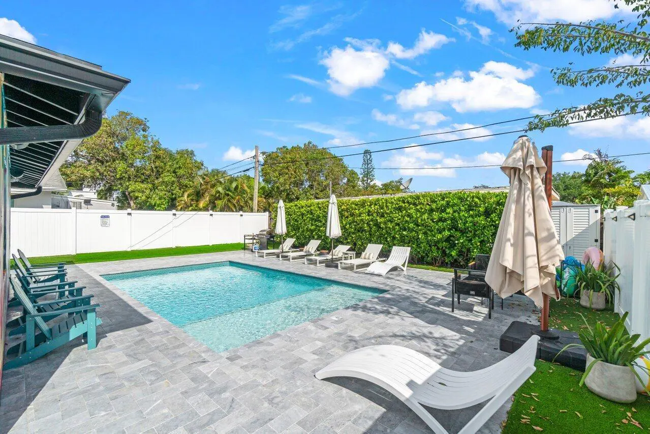A backyard with a pool, lounge chairs, and a white fence under a blue sky with scattered clouds.