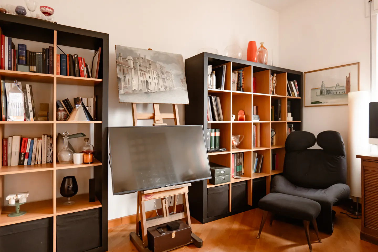 A living room with two black bookshelves filled with books and decorative objects. A TV is on an easel, and a black chair is in the corner.