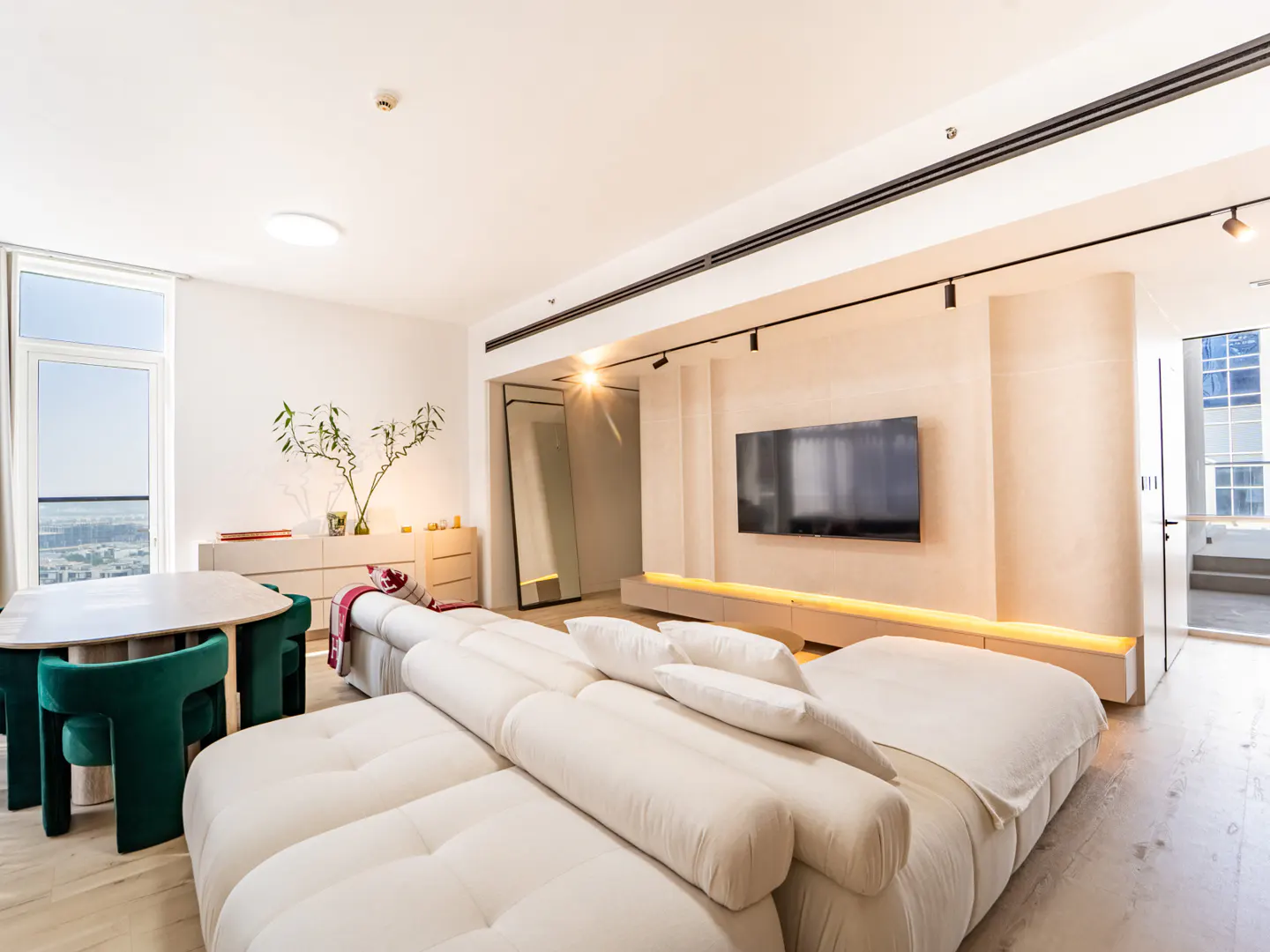 Modern living room with a large white modular sofa, wall-mounted TV, and a dining area with green velvet chairs.