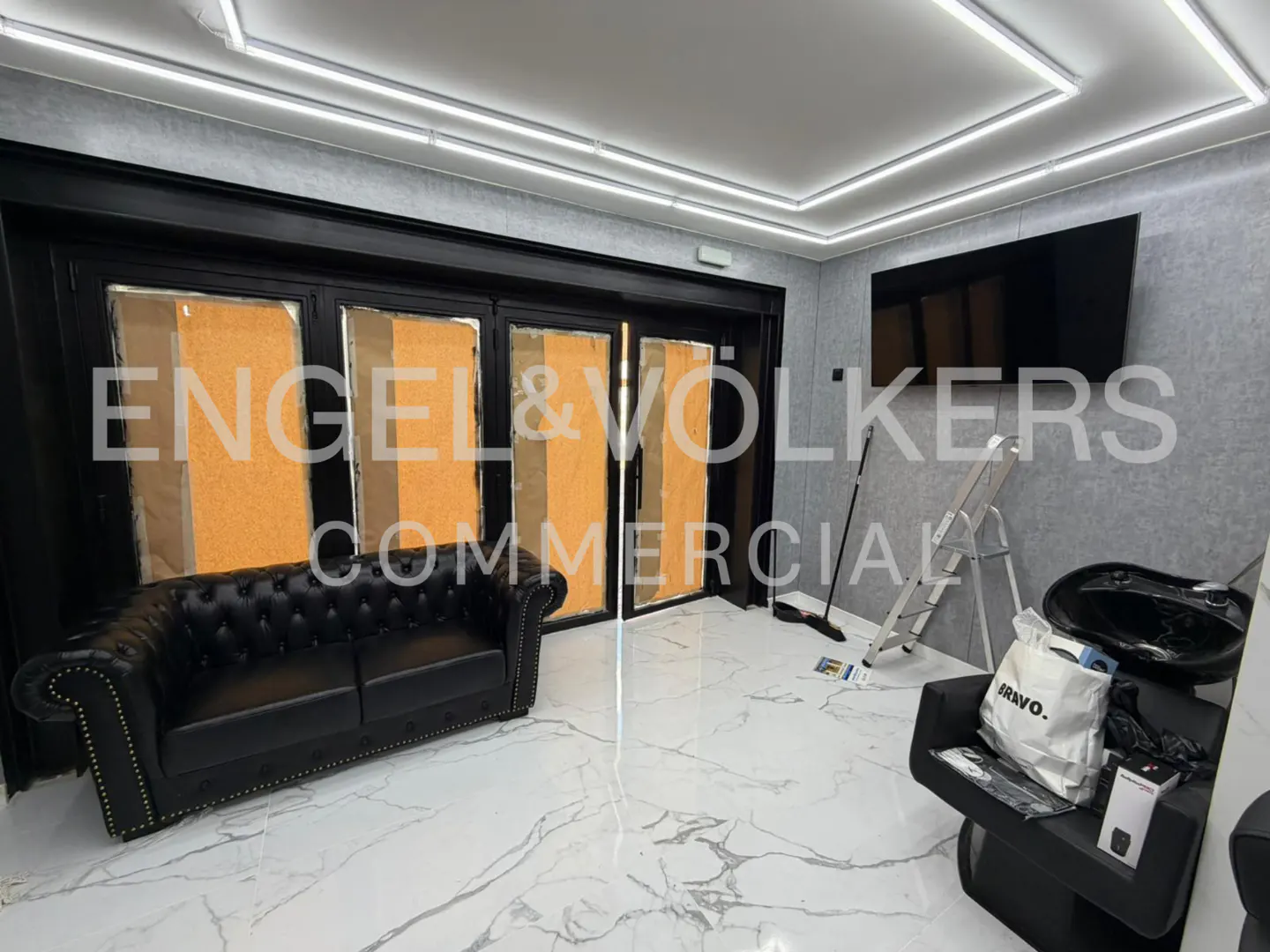 Interior view of a commercial space with a black leather sofa, salon chair, and marble floors. The walls are gray, and there are black-framed windows.