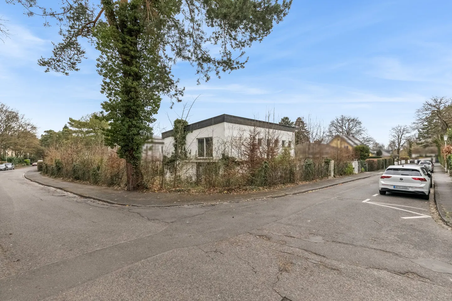 Street view of a modern white house with a flat roof, surrounded by trees and bushes. A white car is parked on the right side of the street.