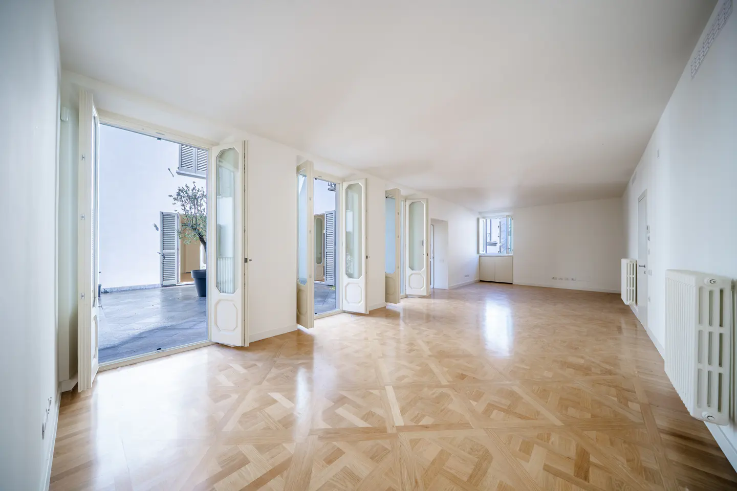Bright, empty room with parquet floors and white walls. French doors open to a patio with a potted tree. Radiators are visible on the right wall.