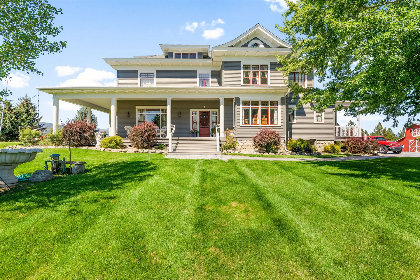 A two-story gray house with a large green lawn and a red barn in the background.