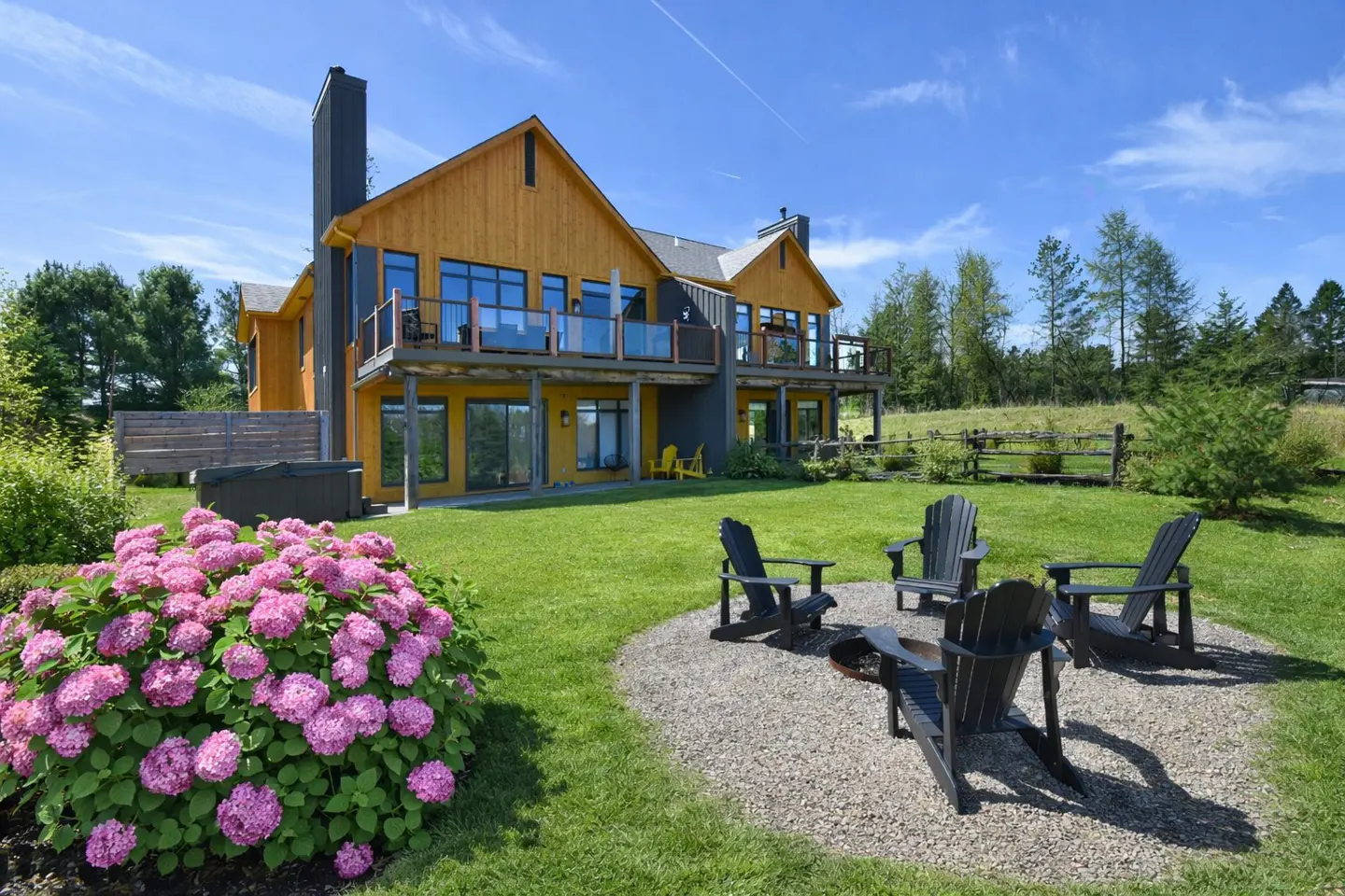 Two-story yellow house with a black chimney, a gravel fire pit with black chairs, and a pink hydrangea bush.