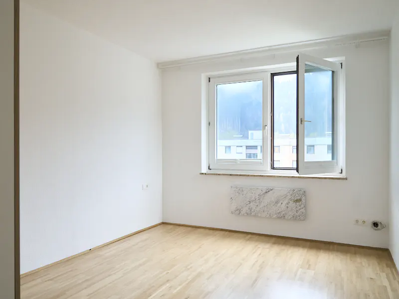 Empty room with light wood floors, white walls, and a large window with a view of buildings and trees.