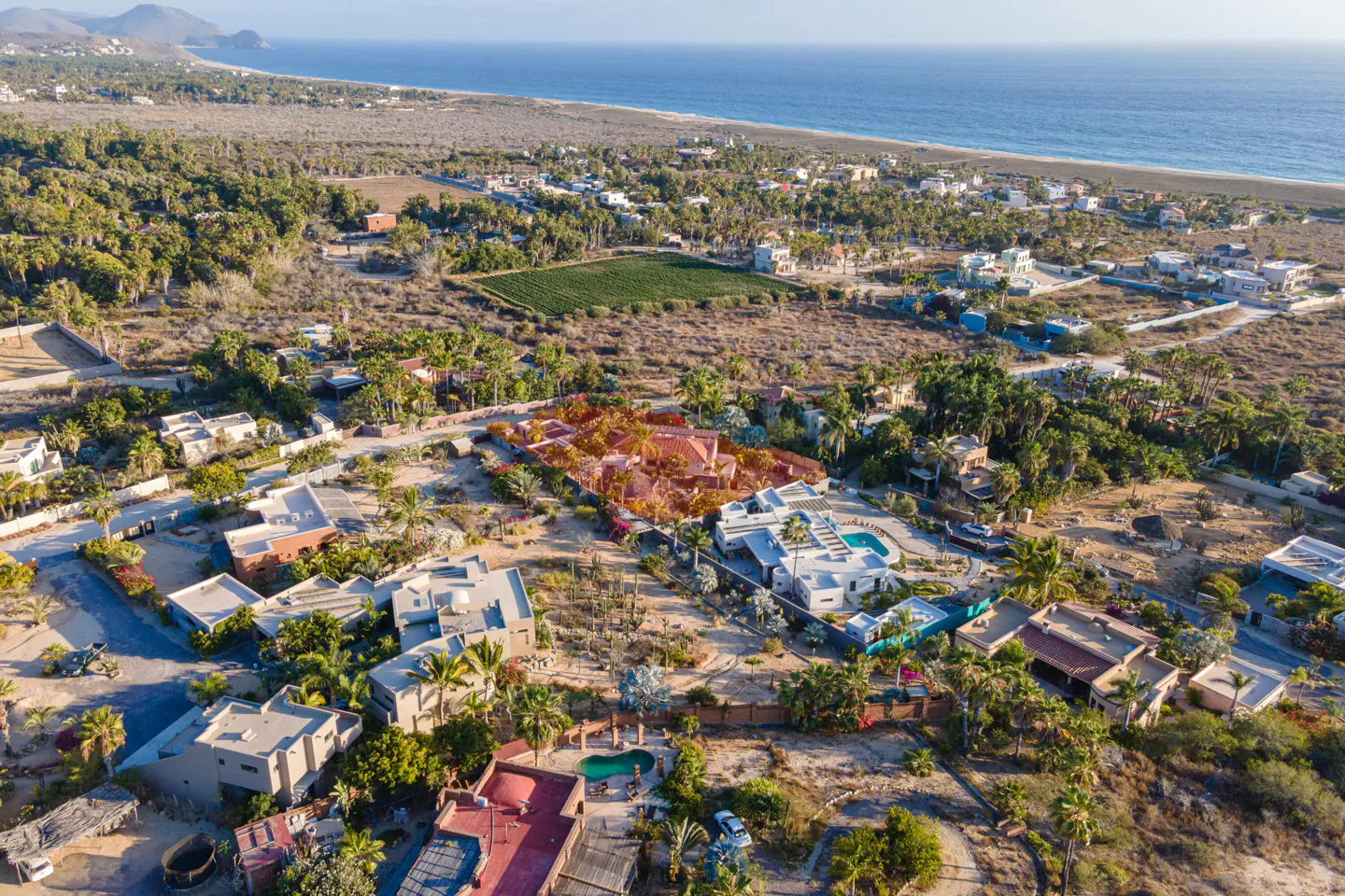 Aerial view of luxury homes with pools, palm trees, and ocean backdrop. Houses are white, red, and tan.