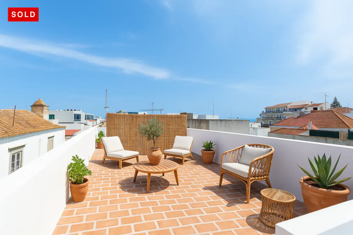 A rooftop patio with terracotta tiles, white walls, and wicker furniture under a blue sky. A "SOLD" sign is in the upper left corner.