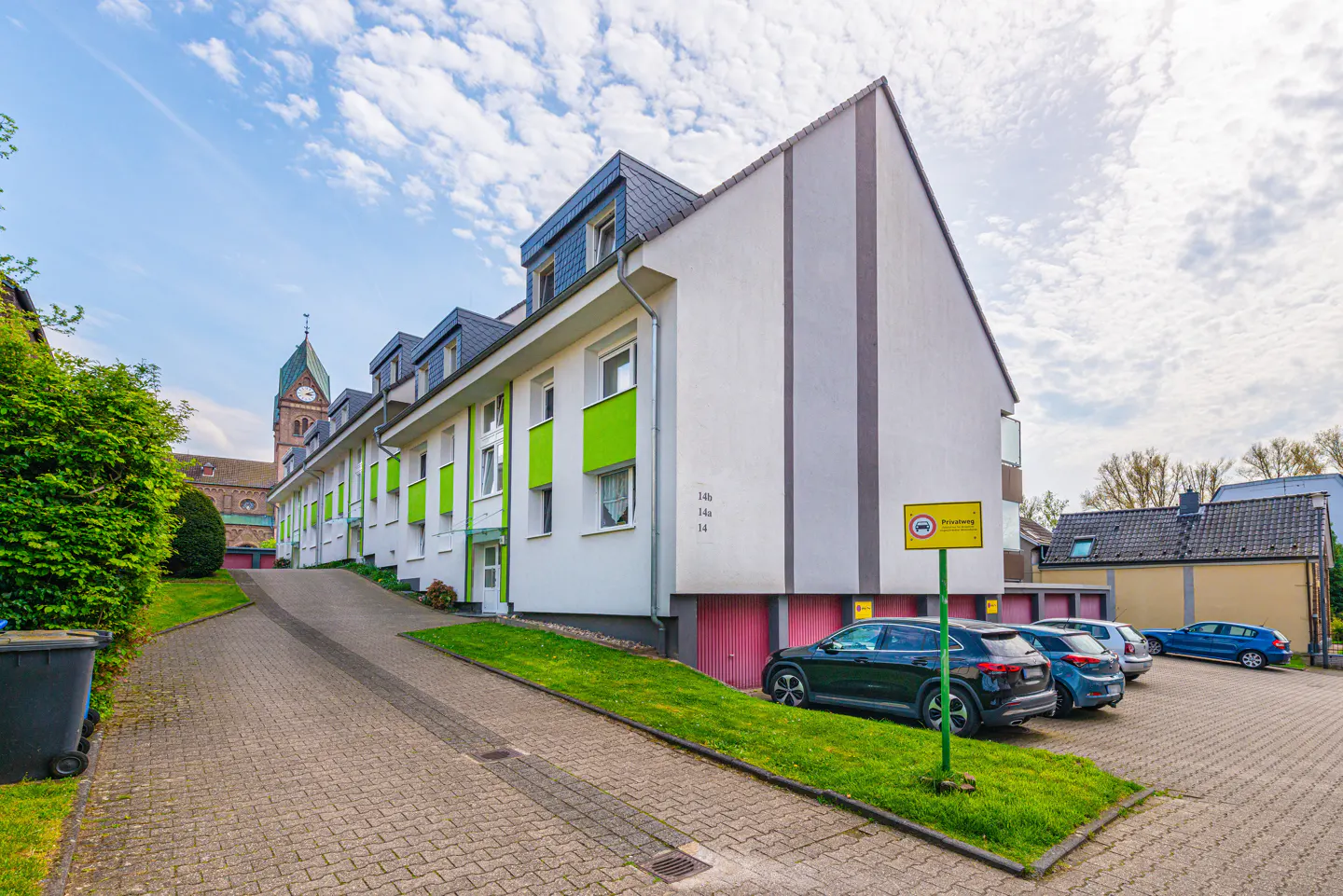 Row of white townhouses with green accents, garages, and parked cars. A church tower is visible in the background.
