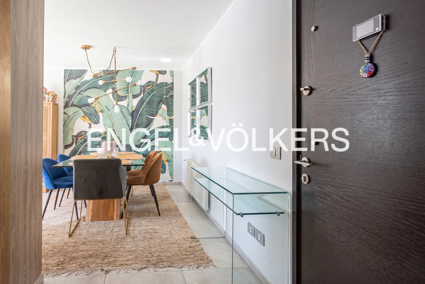 Modern dining room with tropical leaf wallpaper, glass console table, and dark wood door in the foreground.