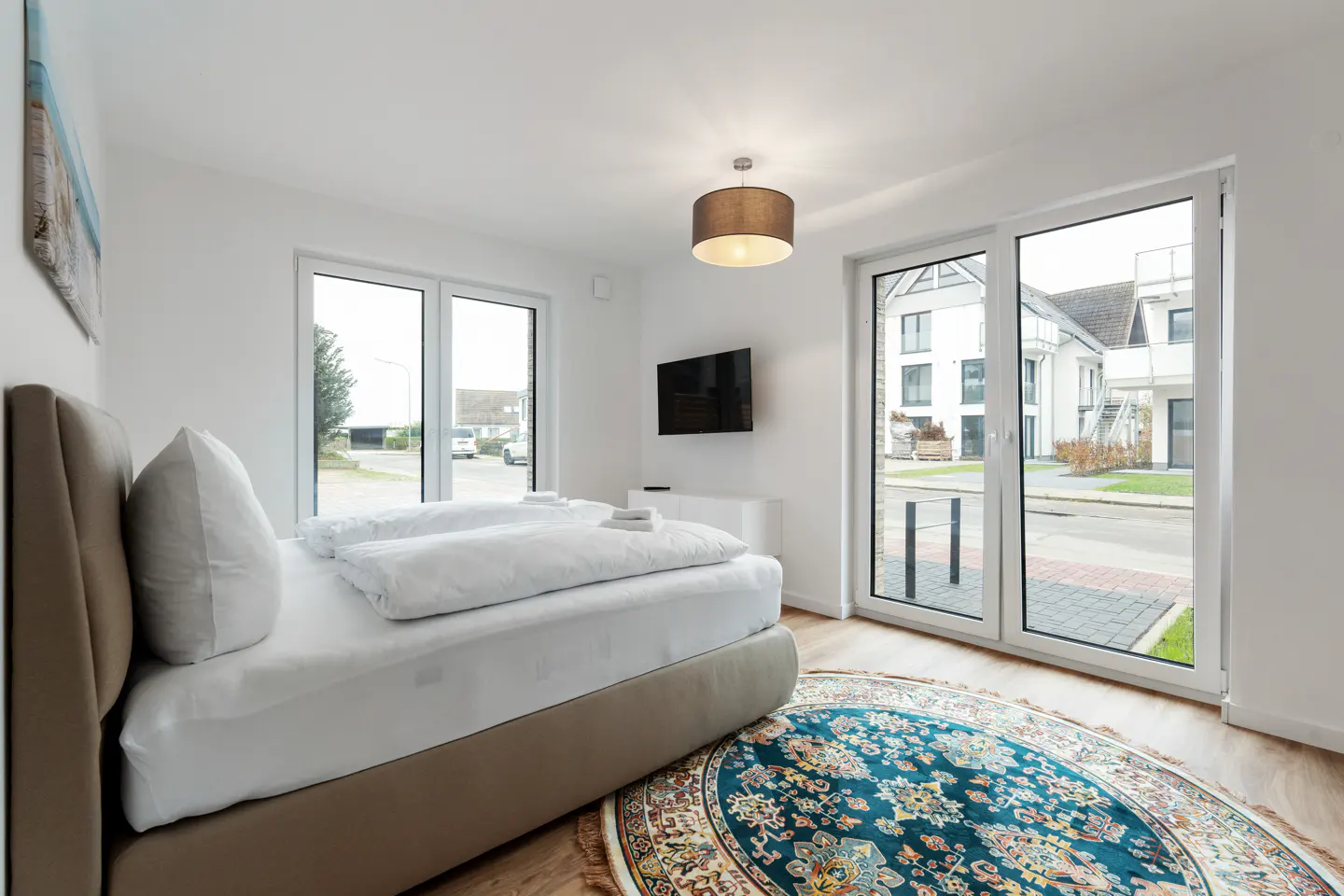 Bright bedroom with a bed, white linens, and a blue floral rug. Large windows offer a view of a modern building across the street. A TV is mounted on the wall.