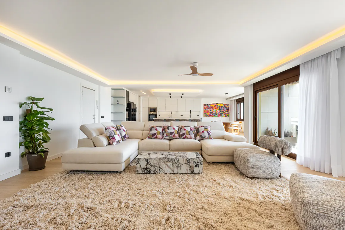 Bright, modern living room with a large white sectional sofa, marble coffee table, and plush beige rug. Kitchen visible in the background.