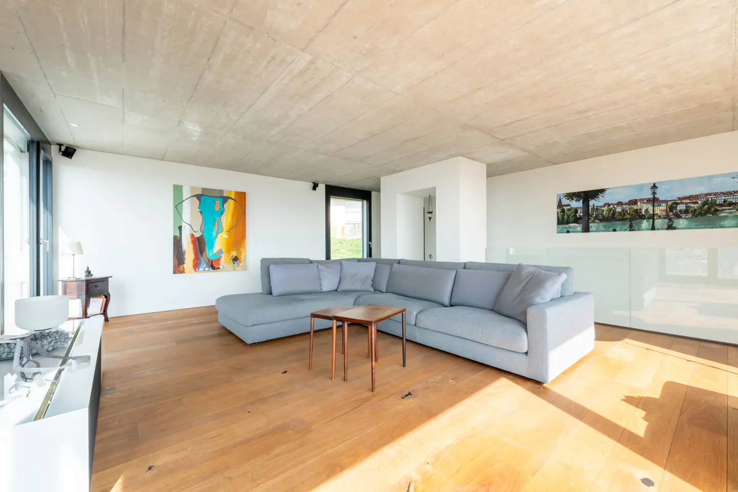 Modern living room with a large grey sectional sofa, wooden floors, concrete ceiling, and colorful abstract wall art.