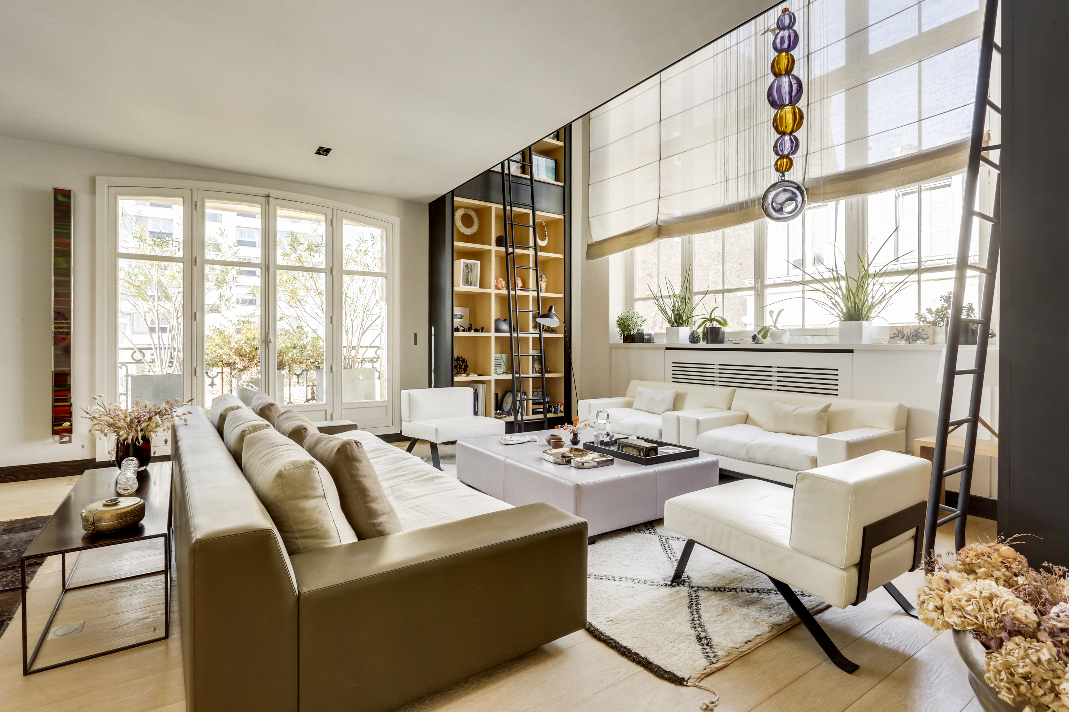 Modern living room with white sofas, a large bookshelf with a ladder, and floor-to-ceiling windows with natural light. [AI-generated text]