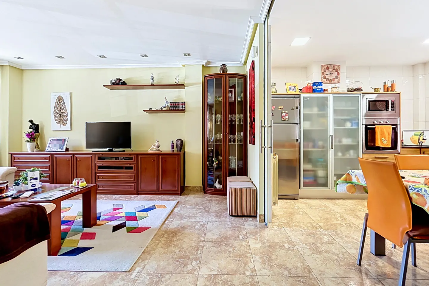 A bright living room with a TV, wooden furniture, and a colorful rug, leading to a kitchen with stainless steel appliances.
