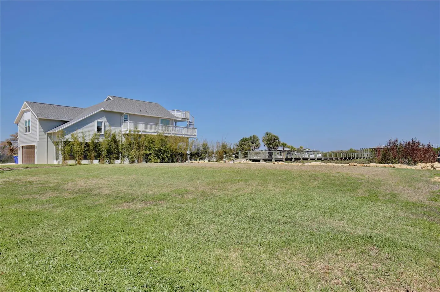 A two-story gray house with a deck and spiral staircase sits on a grassy lawn under a clear blue sky.