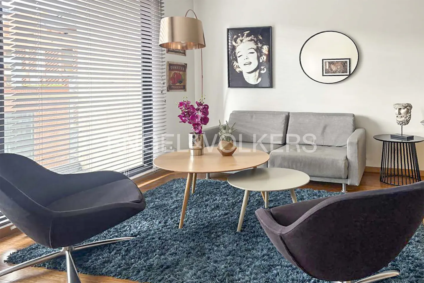 Living room with gray sofa, round wood tables, and two black chairs on a blue rug. A Marilyn Monroe portrait hangs on the wall.