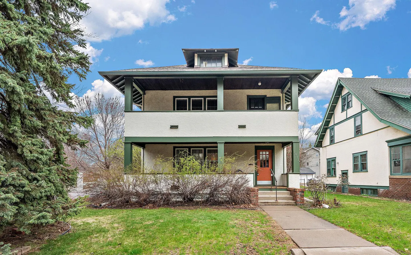 Two-story house with white stucco, green trim, and an orange front door. A lawn and trees are in the foreground.