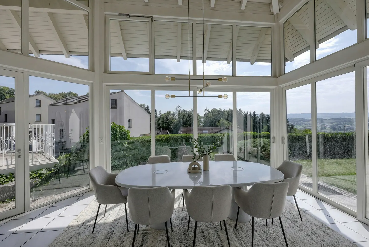 Bright sunroom with white-framed windows, an oval table with gray chairs, and a modern chandelier. Outside, green trees and houses are visible.