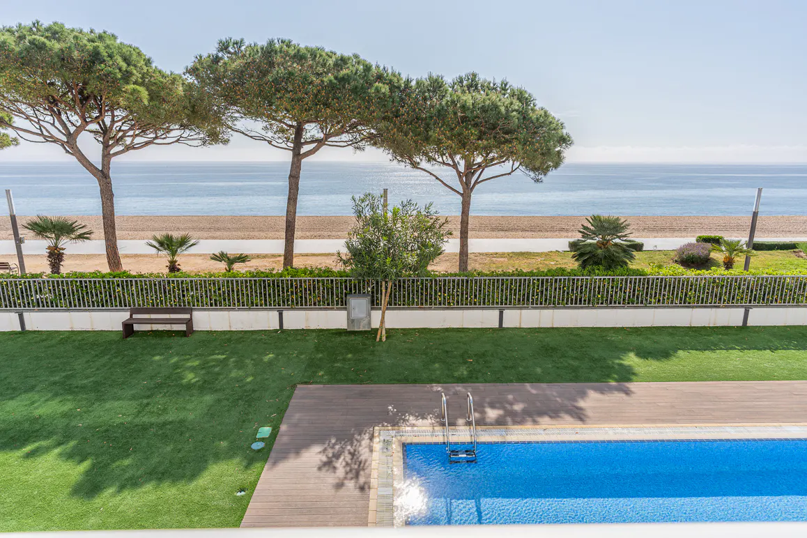 Aerial view of a blue pool with wooden deck, green lawn, and beach with trees in the background.
