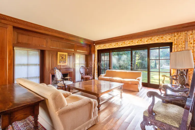 A sunlit living room with wood paneling, hardwood floors, and tan sofas. A large window overlooks a green lawn.
