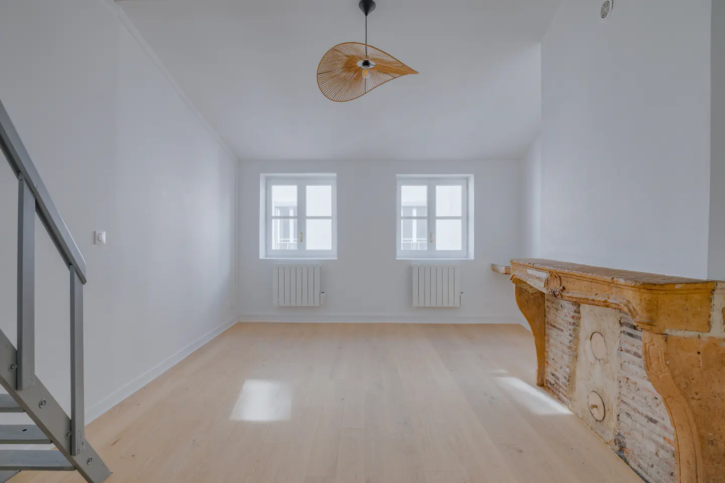 Bright, empty room with white walls, light wood floors, and two windows. A decorative fireplace and a modern light fixture add character.
