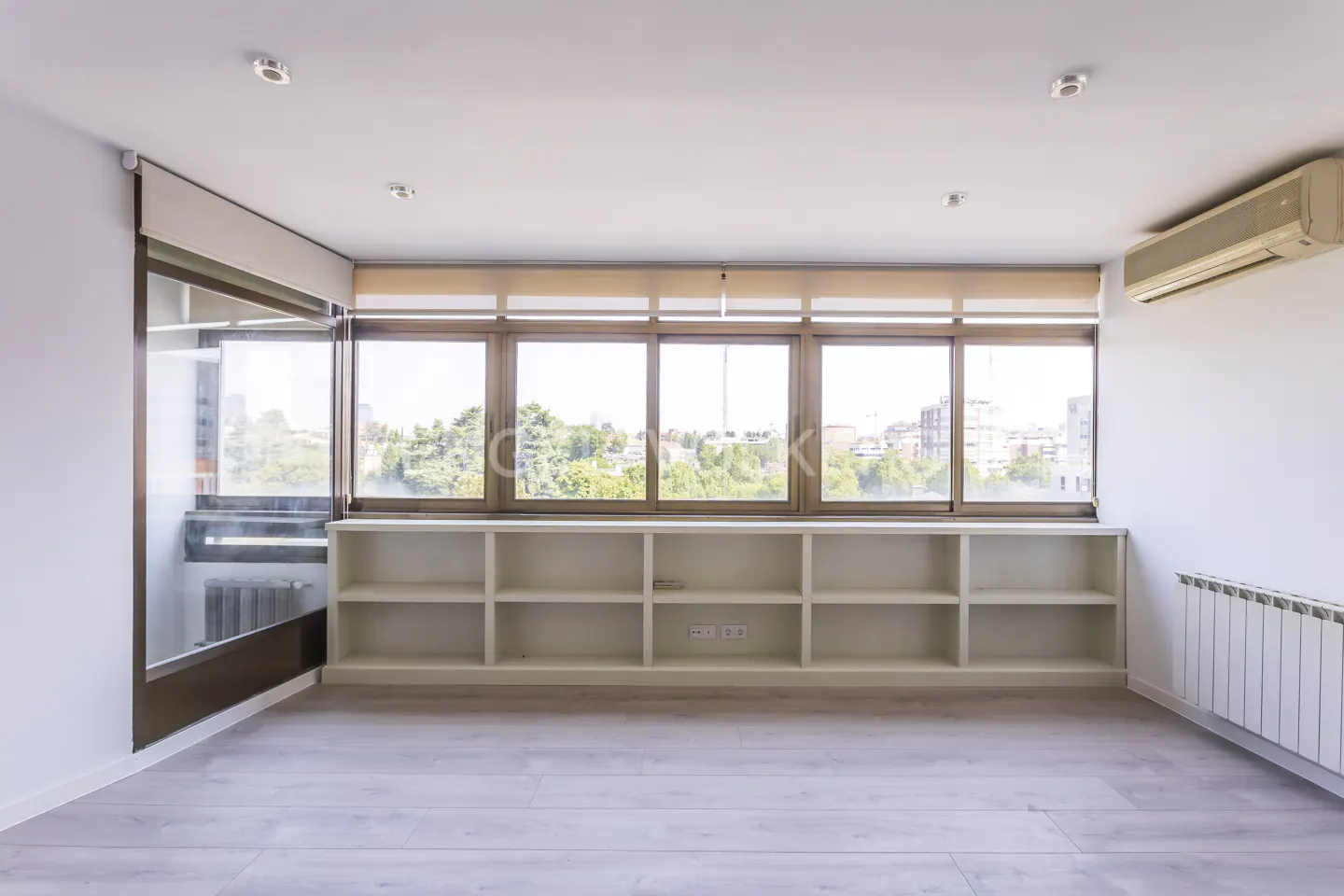 Bright, empty room with light wood floors, white walls, and large windows overlooking trees and buildings. Built-in white shelves line the wall.