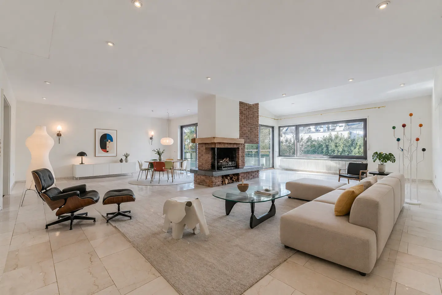 Bright, open living room with a brick fireplace, beige sectional sofa, and Eames chair. Dining table and chairs are in the background.
