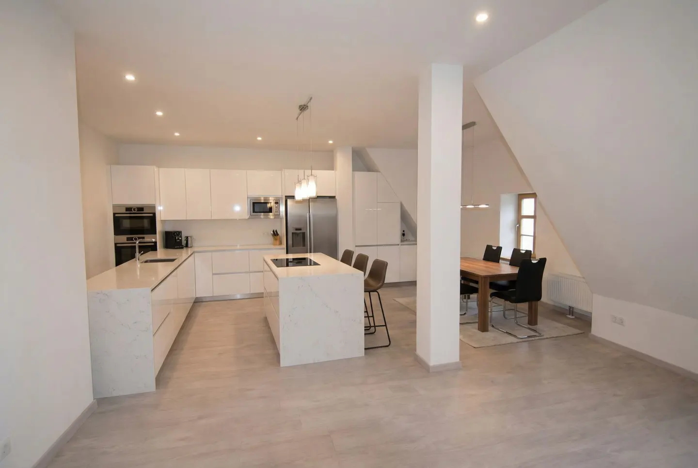 Bright, open kitchen with white cabinets, marble countertops, and island. Dining table with black chairs sits near a window in the background.