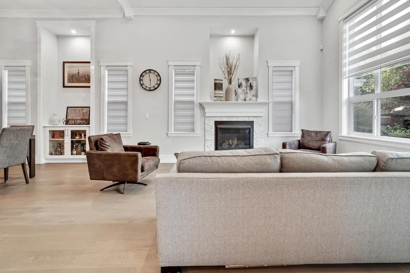 Bright living room with a gray sofa, brown leather chairs, and a white fireplace with a black firebox. Windows with blinds and light wood floors.