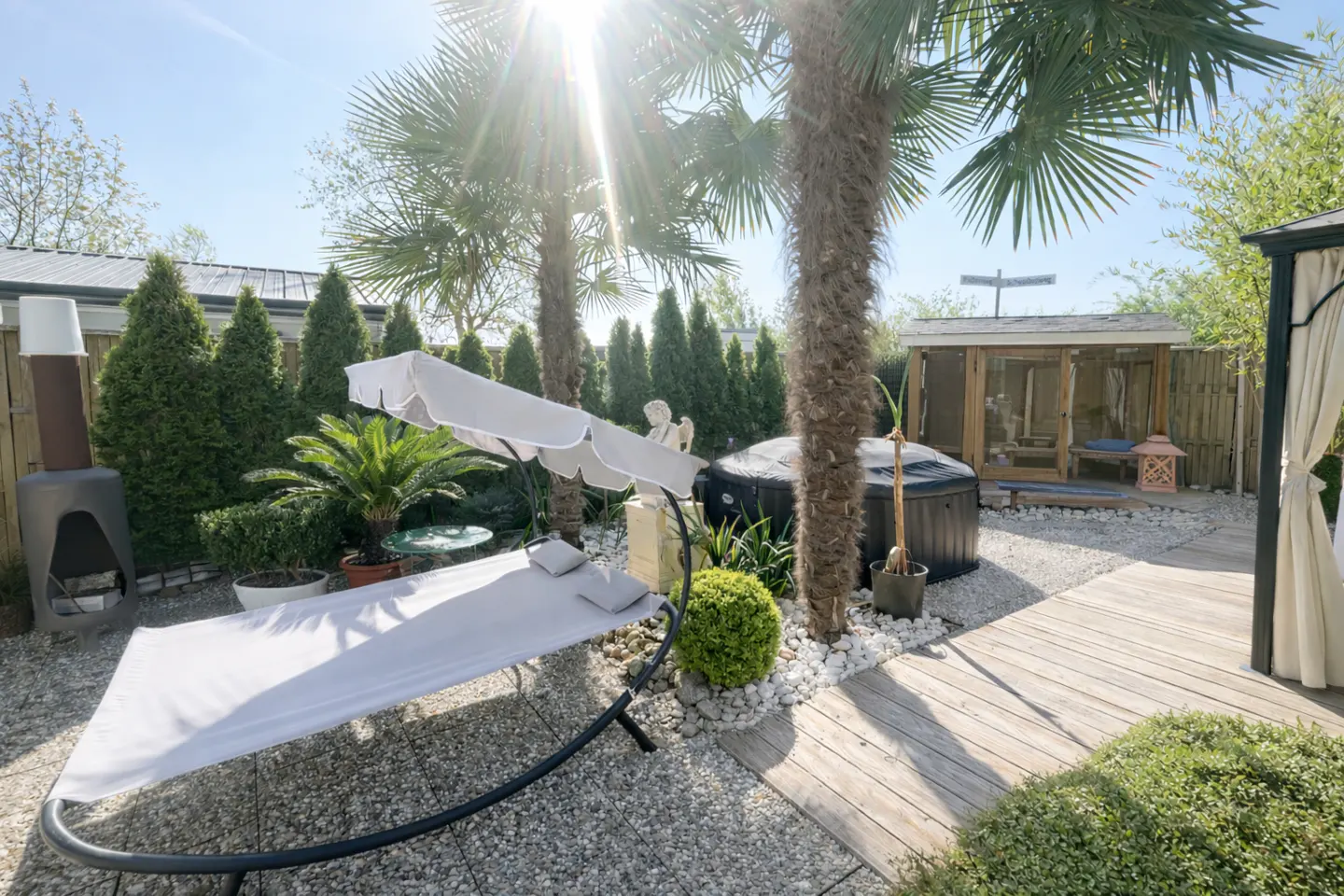 Sunny backyard with a white hammock, hot tub, palm trees, and a wooden gazebo on a gravel and deck patio.