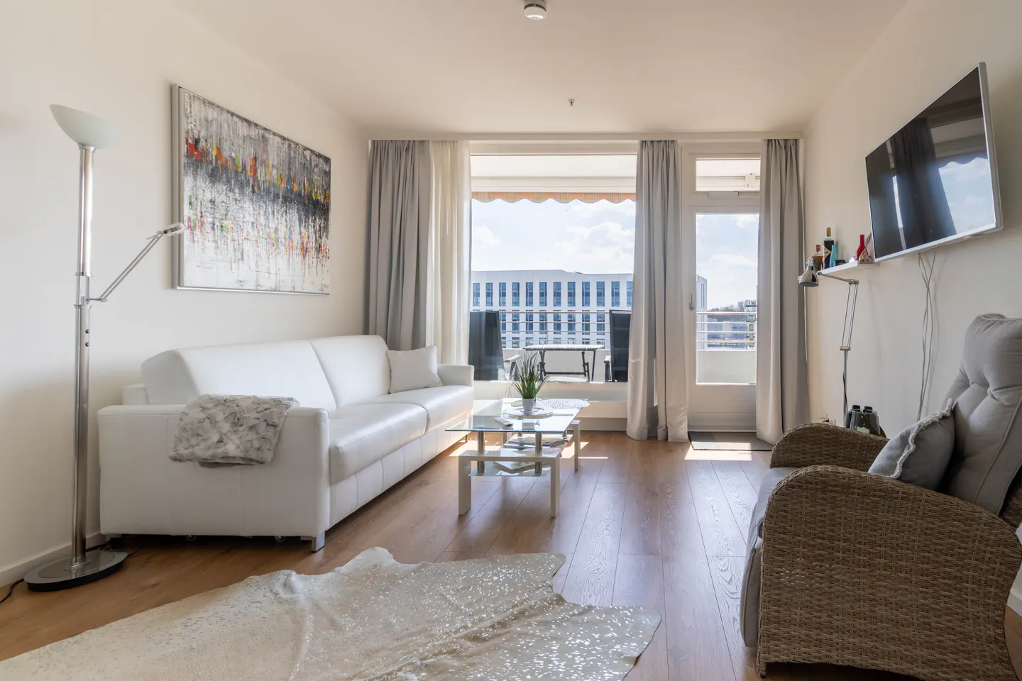 Bright living room with white sofa, glass table, and wicker chair. Abstract art hangs above the sofa, and a balcony overlooks a city.