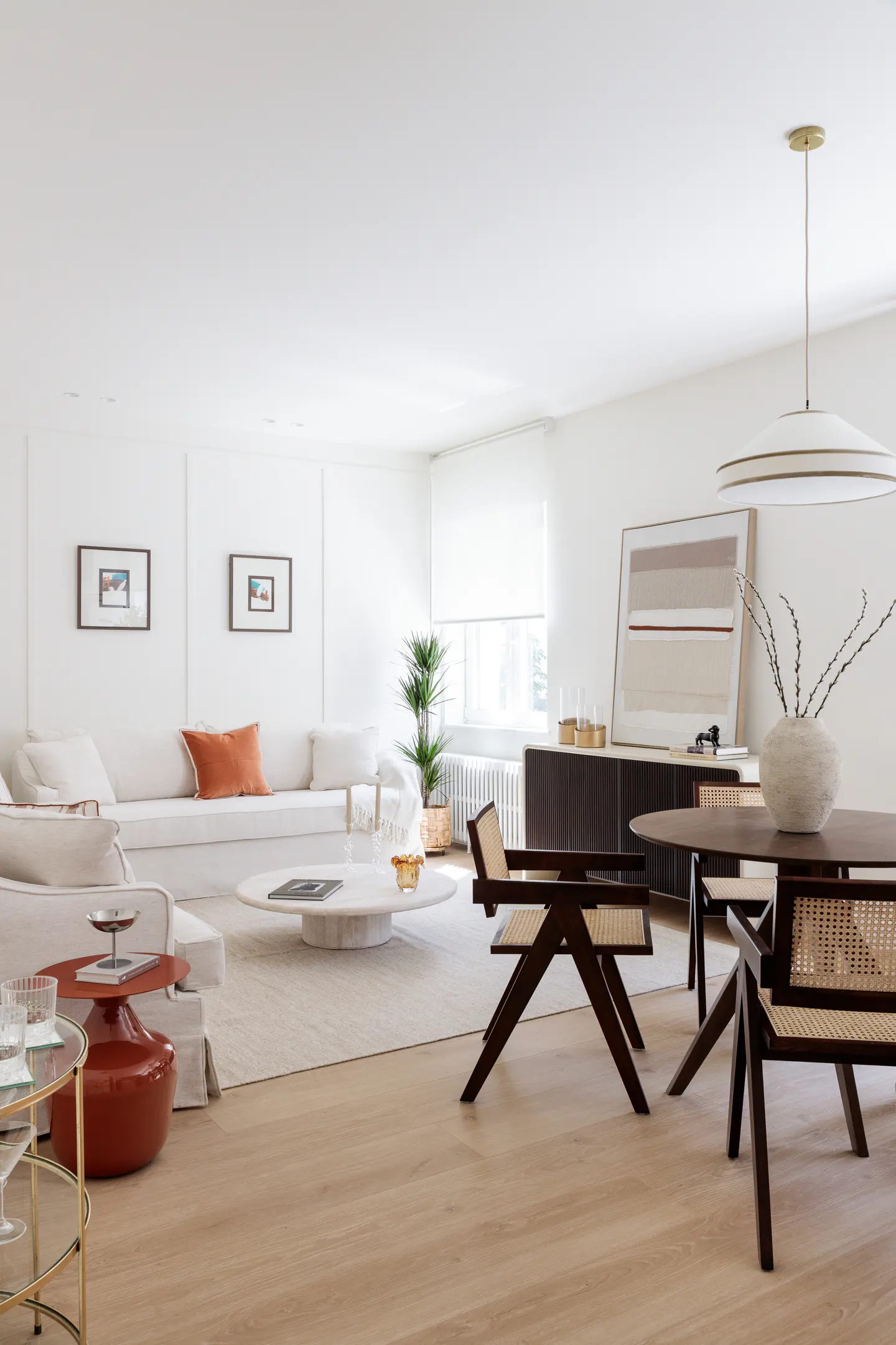 Bright, modern living room with white walls, light wood floors, and neutral furniture. A round table and chairs sit near a large window.