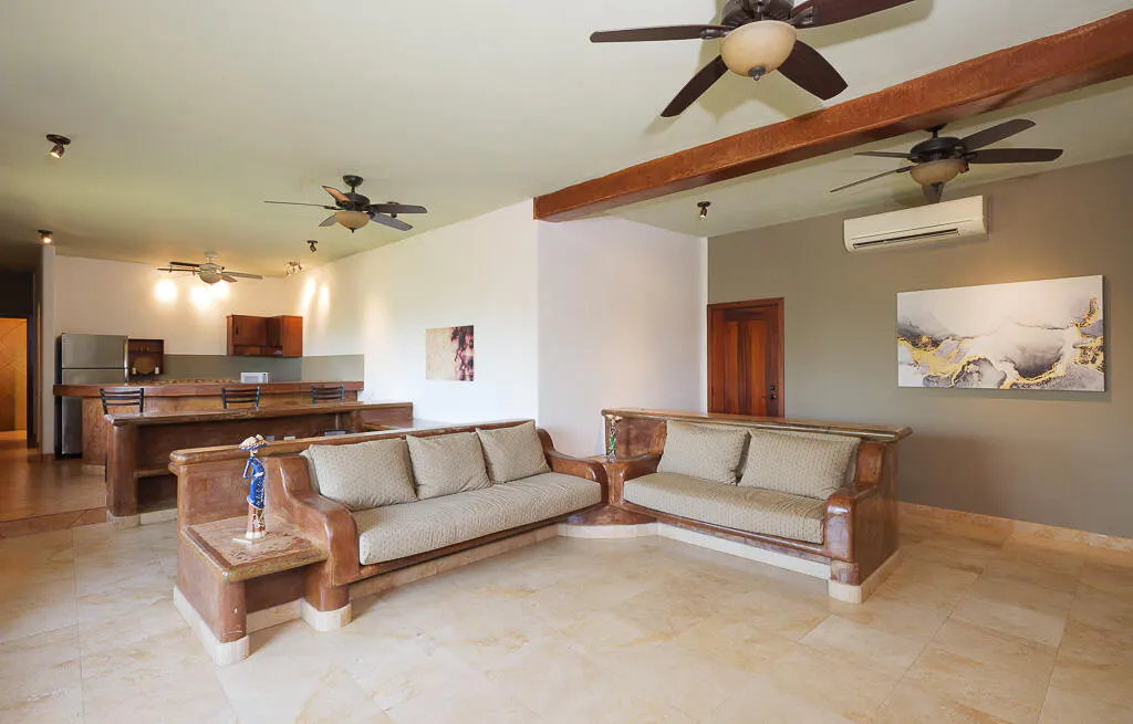 Living room with two sofas, a kitchen, and three ceiling fans. The walls are white and gray, and the floor is tiled.