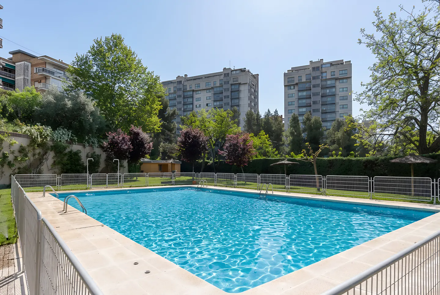 Outdoor pool with turquoise water, surrounded by a fence, trees, and apartment buildings under a clear blue sky.