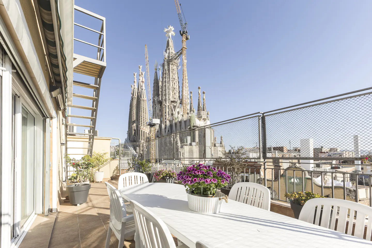 Barcelona rooftop view of Sagrada Familia. White table and chairs on a patio with potted plants. Construction cranes visible.