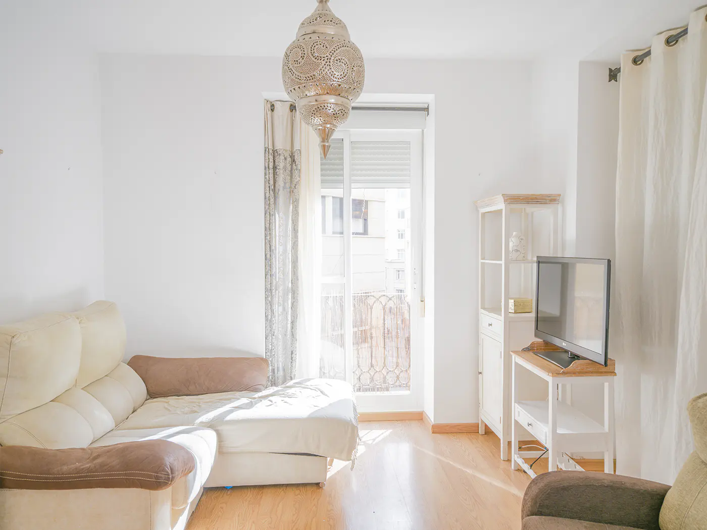 Bright living room with a beige sectional sofa, a white cabinet, and a TV. A decorative lamp hangs from the ceiling. Balcony door lets in natural light.
