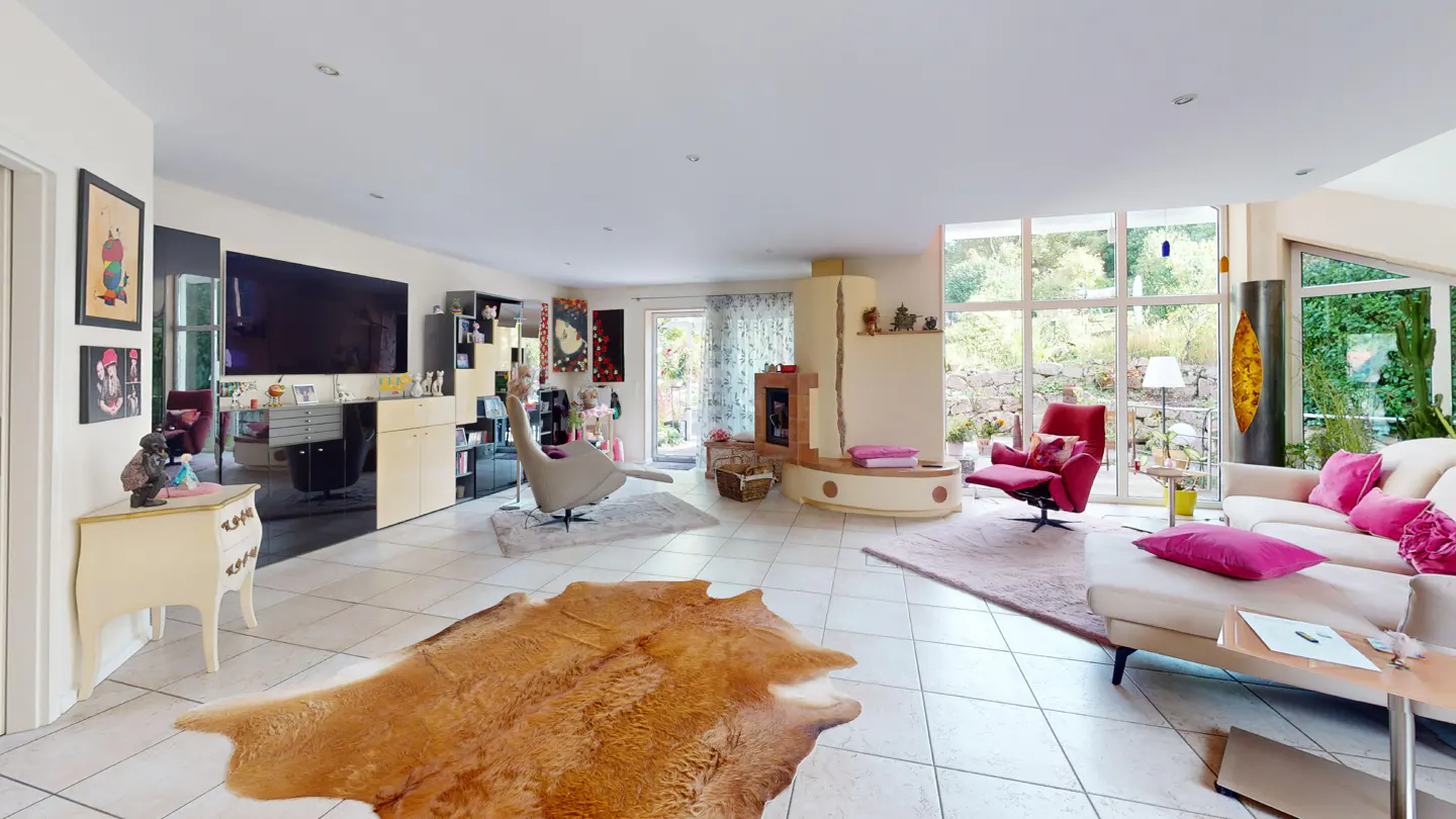 Bright living room with white tile floor, a brown hide rug, a fireplace, and a white sofa with pink pillows. Large windows overlook a garden.