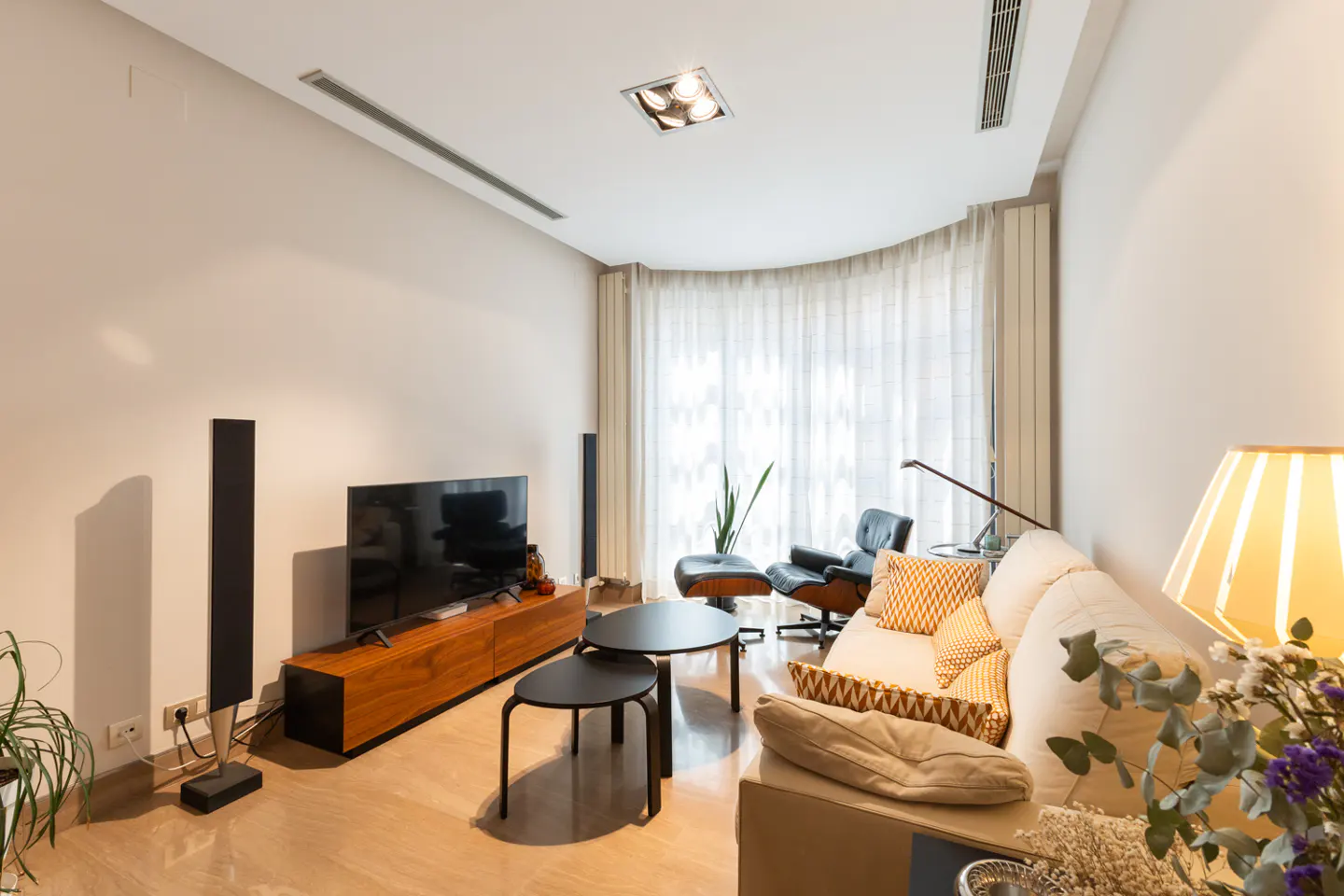 Bright living room with a cream sofa, black speakers, and a wooden TV stand. A black chair sits near sheer curtains.